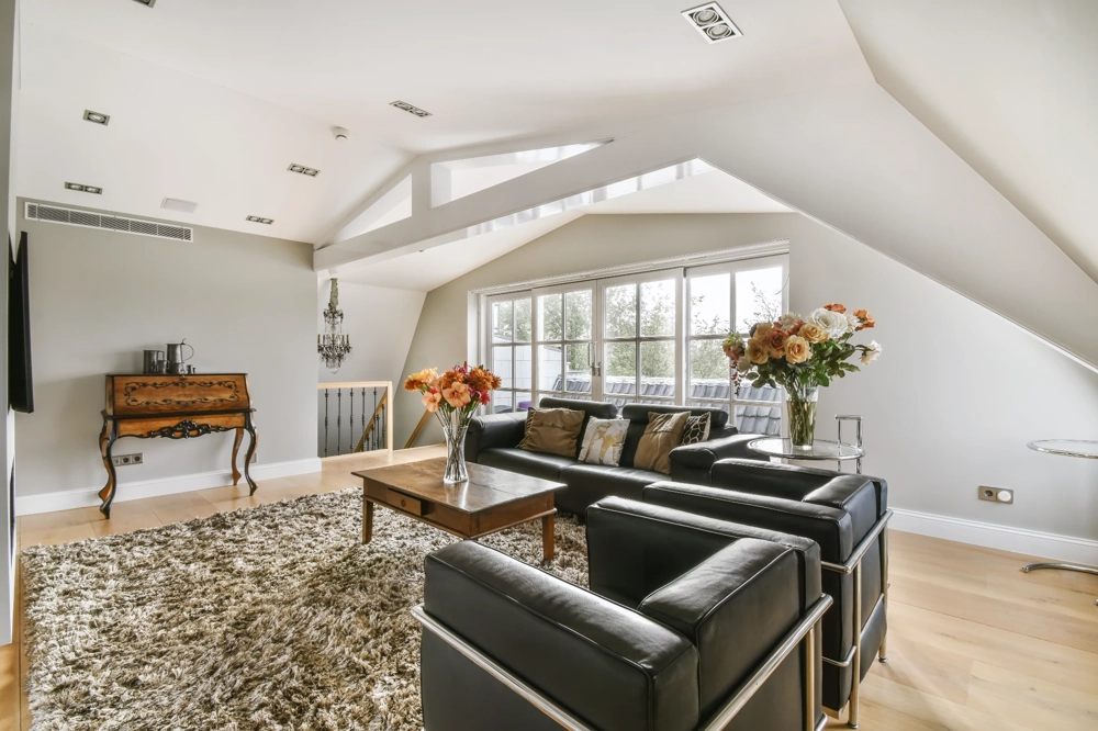 Przestronny salon w adaptowanym poddaszu z czarnymi skórzanymi sofami, drewnianym stolikiem, dywanem i dekoracyjnymi kwiatami Adaptacja poddasza