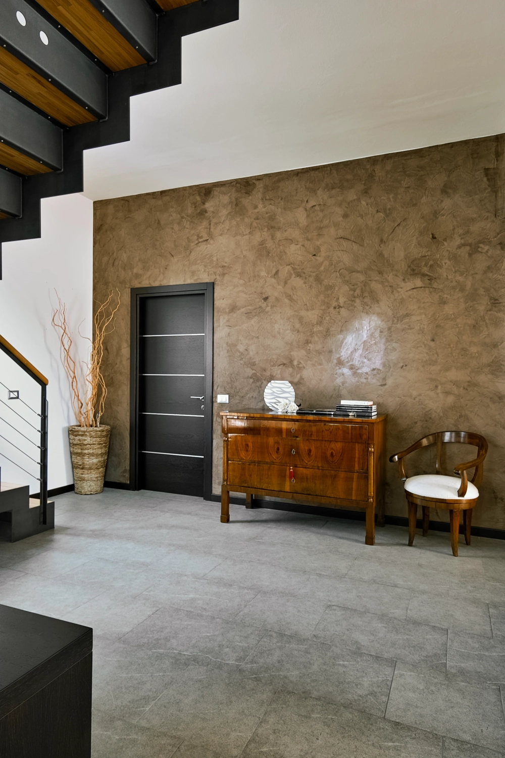 Nowoczesny korytarz z szarą podłogą z płytek, czarnymi schodami i drewnianą komodą – Układanie płytek