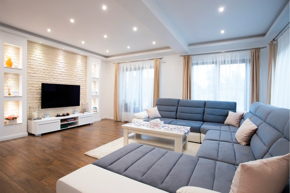 Salon z szarym narożnikiem, białym stolikiem, drewnianą podłogą i sufitami podwieszanymi z oświetleniem LED